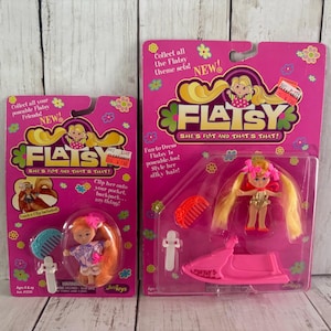 May include: Two Flatsy dolls in original packaging. The pink packaging features the Flatsy logo, a cartoon girl, and the text "She's Flat and That's That!". One doll includes a comb and clip, the other a pink jet ski.