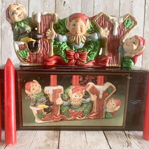 May include: A ceramic Christmas candle holder featuring the word "JOY" with elves holding candles. The holder is decorated with holly, red bows, and green leaves. Four red candles are included.