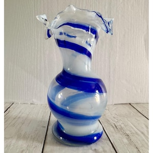 May include: A decorative glass vase with a white and blue swirl design. The vase has a bulbous body, a slender neck, and a ruffled rim. The blue stripes are a vibrant contrast against the white glass. The vase sits on a wooden surface.