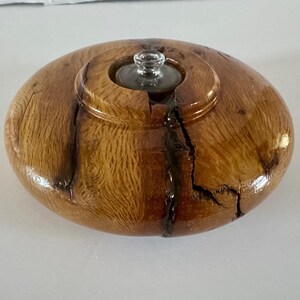 May include: Oval wooden container with a clear glass lid. The container is made of light brown wood with dark brown cracks and knots. The glass lid is small and round, with a decorative top.