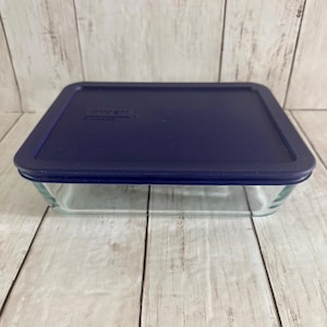 May include: A rectangular clear glass food storage container with a dark blue plastic lid. The lid features a flat top surface with a slightly raised border and the embossed text Pyrex visible on the upper left corner of the lid surface.