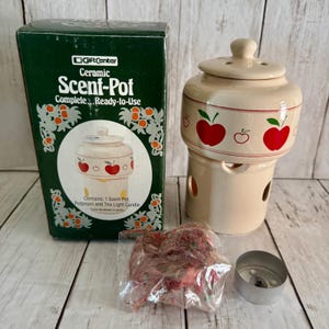 May include: A cream-colored ceramic scent pot with a lid, featuring a red apple pattern. The set includes a dark green cardboard box labeled GiftCenter Ceramic Scent-Pot, a clear plastic bag of dried potpourri, and a small metal tea light candle.