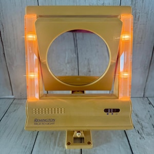 May include: A vintage Remington True-To-Light vanity mirror with a yellow plastic frame. The square mirror features two vertical side panels with integrated glowing orange lights. A sliding power switch is located on the base below the mirror.