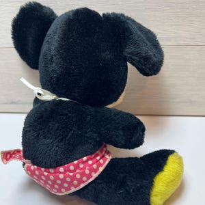 K&ouml;nnte beinhalten: Ein schwarzes Pl&uuml;schmaus-Spielzeug mit gro&szlig;en Ohren, einem wei&szlig;en Bauch und einer rosa-wei&szlig; gepunkteten Sch&auml;rpe. Die F&uuml;&szlig;e der Maus sind gelb. Das Spielzeug liegt auf einer wei&szlig;en Oberfl&auml;che.