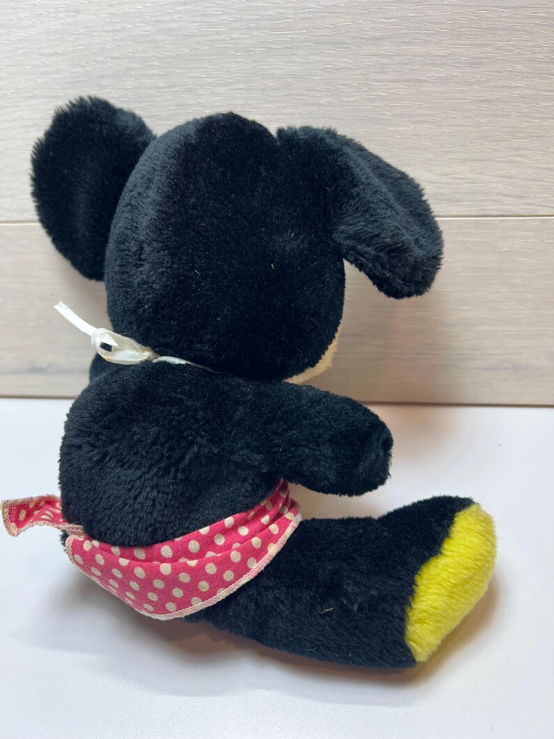 K&ouml;nnte beinhalten: Ein schwarzes Pl&uuml;schmaus-Spielzeug mit gro&szlig;en Ohren, einem wei&szlig;en Bauch und einer rosa-wei&szlig; gepunkteten Sch&auml;rpe. Die F&uuml;&szlig;e der Maus sind gelb. Das Spielzeug liegt auf einer wei&szlig;en Oberfl&auml;che.