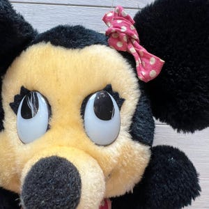 K&ouml;nnte beinhalten: Nahaufnahme eines Vintage-Pl&uuml;schtiers mit einer Zeichentrickfigur. Das Spielzeug hat eine schwarze Nase, schwarze Ohren und ein gelbes Gesicht. Eine rosa Schleife mit wei&szlig;en Punkten ziert den Kopf. Das Spielzeug tr&auml;gt ein rosa Kleid mit wei&szlig;en Punkten.