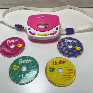 Peut inclure: Un lecteur CD Barbie rose et blanc avec une sangle blanche et quatre CD colorés. Les CD sont roses, violets, verts et jaunes, chacun avec le logo Barbie et des titres de chansons. Le lecteur CD a un affichage numérique.