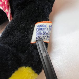 K&ouml;nnte beinhalten: Nahaufnahme eines schwarzen Pl&uuml;schtiers mit gelben und rosa Akzenten. Ein Etikett mit Text ist am Spielzeug befestigt. Auf dem Etikett steht "MINNIE MOUSE PRODUCT OF...SHREDDED...GROUND NU...WALT DIS..."