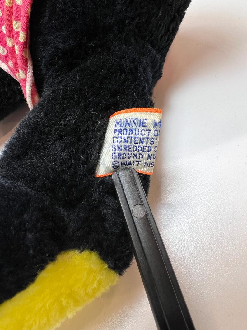 K&ouml;nnte beinhalten: Nahaufnahme eines schwarzen Pl&uuml;schtiers mit gelben und rosa Akzenten. Ein Etikett mit Text ist am Spielzeug befestigt. Auf dem Etikett steht "MINNIE MOUSE PRODUCT OF...SHREDDED...GROUND NU...WALT DIS..."
