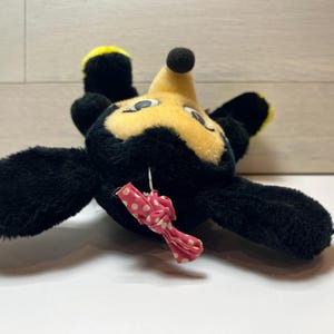 K&ouml;nnte beinhalten: Ein Vintage-Pl&uuml;schtier in Form einer Zeichentrickfigur mit schwarzem Fell, gelben Akzenten und einem gelben Gesicht. Es hat eine kleine schwarze Nase und eine rosa Schleife mit wei&szlig;en Punkten. Das Spielzeug liegt auf dem R&uuml;cken.