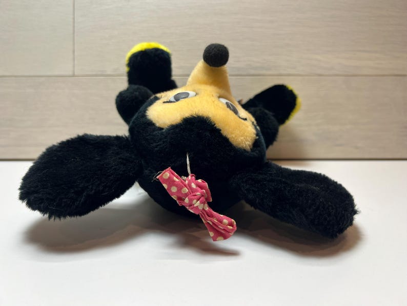 K&ouml;nnte beinhalten: Ein Vintage-Pl&uuml;schtier in Form einer Zeichentrickfigur mit schwarzem Fell, gelben Akzenten und einem gelben Gesicht. Es hat eine kleine schwarze Nase und eine rosa Schleife mit wei&szlig;en Punkten. Das Spielzeug liegt auf dem R&uuml;cken.