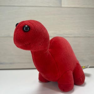 May include: A plush red dinosaur toy with black button eyes. The stuffed animal has a rounded head, a long neck, and a small body with four stubby legs. The toy is made of a soft, fuzzy material.