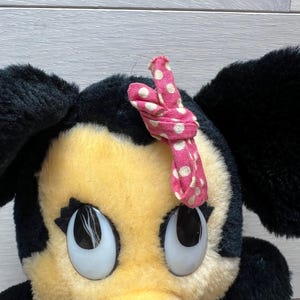 K&ouml;nnte beinhalten: Ein Pl&uuml;schtier mit einem Zeichentrickcharakter mit gro&szlig;en, ausdrucksstarken Augen. Die Figur hat schwarze Ohren, ein gelbes Gesicht und eine rosa Schleife mit wei&szlig;en Punkten. Das Spielzeug ist ein klassisches Design.