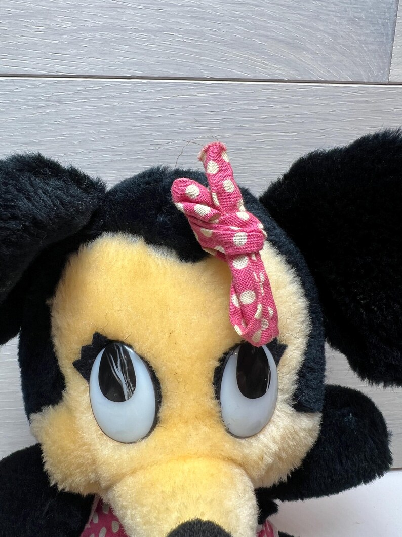 K&ouml;nnte beinhalten: Ein Pl&uuml;schtier mit einem Zeichentrickcharakter mit gro&szlig;en, ausdrucksstarken Augen. Die Figur hat schwarze Ohren, ein gelbes Gesicht und eine rosa Schleife mit wei&szlig;en Punkten. Das Spielzeug ist ein klassisches Design.