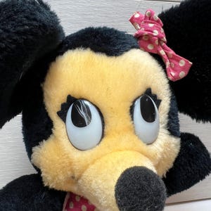 K&ouml;nnte beinhalten: Ein Vintage-Pl&uuml;schtier mit einer Zeichentrickfigur mit schwarzem Kopf und Ohren, einem gelben Gesicht und gro&szlig;en Augen. Das Spielzeug hat eine rosa Schleife mit Punkten und ein passendes Kleid. Ein klassisches Sammlerst&uuml;ck f&uuml;r Kinder.