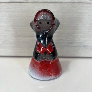 May include: A ceramic figurine of a woman wearing a red and black dress and a black hooded cloak. The figurine has a white glaze and a distressed look.