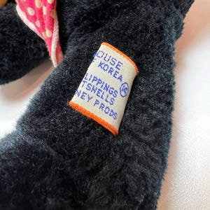K&ouml;nnte beinhalten: Nahaufnahme eines schwarzen Pl&uuml;schtiers mit einer rosa Schleife mit Punkten und einem gelben Fu&szlig;. Ein Etikett mit Text ist auf den Arm des Spielzeugs gen&auml;ht. Das Etikett hat einen orangefarbenen Rand und lautet "HOUSE KOREA LIPPINGS TSHELLS NEY PRODS".
