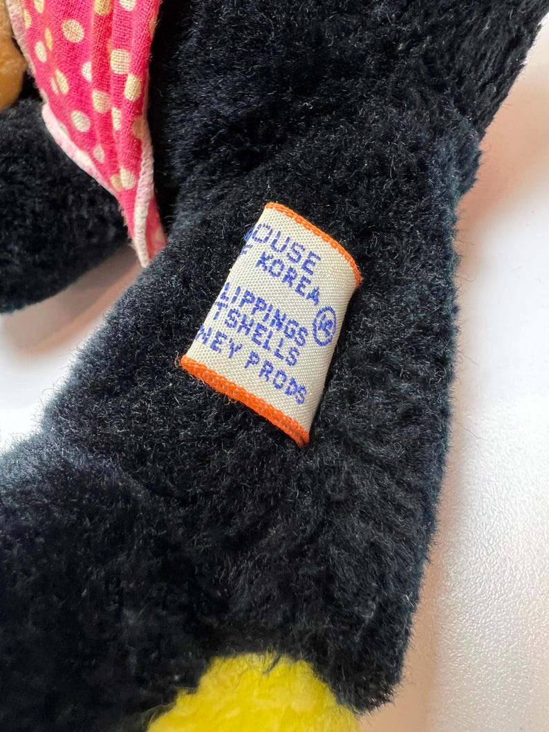 K&ouml;nnte beinhalten: Nahaufnahme eines schwarzen Pl&uuml;schtiers mit einer rosa Schleife mit Punkten und einem gelben Fu&szlig;. Ein Etikett mit Text ist auf den Arm des Spielzeugs gen&auml;ht. Das Etikett hat einen orangefarbenen Rand und lautet "HOUSE KOREA LIPPINGS TSHELLS NEY PRODS".