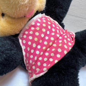 K&ouml;nnte beinhalten: Ein Vintage-Pl&uuml;schtier mit schwarzem K&ouml;rper, beigen Ohren und einem rosa Gesicht. Das Spielzeug tr&auml;gt ein rosa Kleid mit Polka-Dots. Das Pl&uuml;schtier ist weich und scheint sehr geliebt worden zu sein.