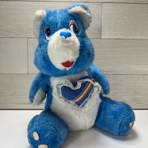 Peut inclure: Une peluche Care Bear bleue avec des détails blancs sur le visage, les oreilles et le ventre. L'ours a un emblème en forme de cœur sur le ventre avec un arc-en-ciel et un cœur bleu. L'ours est assis.
