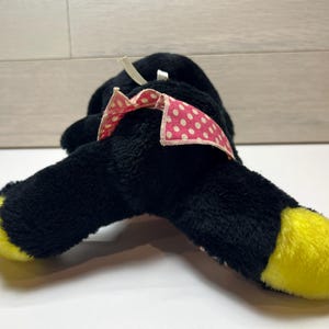 K&ouml;nnte beinhalten: Ein schwarzes Pl&uuml;schtier mit gelben F&uuml;&szlig;en und einer rosa Schleife mit wei&szlig;en Punkten. Das Spielzeug liegt auf der Seite und zeigt sein weiches, strukturiertes Material. Die Schleife f&uuml;gt einen Hauch von Farbe und Detail hinzu.