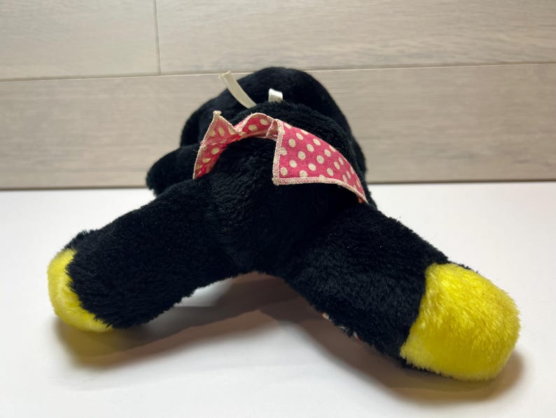 K&ouml;nnte beinhalten: Ein schwarzes Pl&uuml;schtier mit gelben F&uuml;&szlig;en und einer rosa Schleife mit wei&szlig;en Punkten. Das Spielzeug liegt auf der Seite und zeigt sein weiches, strukturiertes Material. Die Schleife f&uuml;gt einen Hauch von Farbe und Detail hinzu.