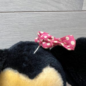 K&ouml;nnte beinhalten: Ein schwarzer Pl&uuml;schtierkopf mit gelbem Gesicht und einer rosa Schleife mit wei&szlig;en Punkten. Die Schleife ist mit wei&szlig;em Faden befestigt. Das Spielzeug hat eine weiche, flauschige Textur.