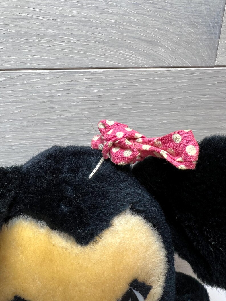 K&ouml;nnte beinhalten: Ein schwarzer Pl&uuml;schtierkopf mit gelbem Gesicht und einer rosa Schleife mit wei&szlig;en Punkten. Die Schleife ist mit wei&szlig;em Faden befestigt. Das Spielzeug hat eine weiche, flauschige Textur.