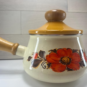 May include: Cream-colored enamelware pot with a lid and handle. The pot features a large orange floral design with brown and green accents. The lid and handle are made of wood.