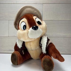 May include: A plush Chipmunk character wearing a brown hat and jacket with white trim. The stuffed animal has a tan face and chest, brown fur, and large black eyes. The toy is sitting on a white surface.