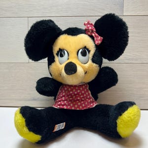 K&ouml;nnte beinhalten: Eine Pl&uuml;schpuppe von Minnie Mouse mit schwarzem Fell, gelben F&uuml;&szlig;en und einem roten Kleid und einer Schleife mit Tupfen. Die Puppe hat gro&szlig;e, ausdrucksstarke Augen und eine schwarze Nase. Die Puppe sitzt mit gespreizten Beinen.