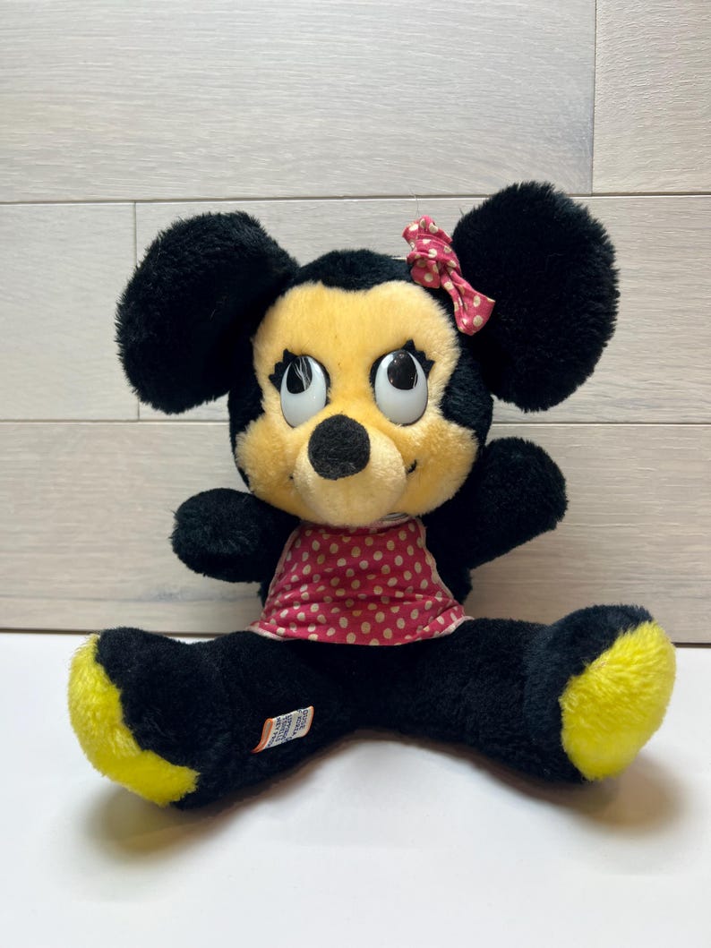 K&ouml;nnte beinhalten: Eine Pl&uuml;schpuppe von Minnie Mouse mit schwarzem Fell, gelben F&uuml;&szlig;en und einem roten Kleid und einer Schleife mit Tupfen. Die Puppe hat gro&szlig;e, ausdrucksstarke Augen und eine schwarze Nase. Die Puppe sitzt mit gespreizten Beinen.