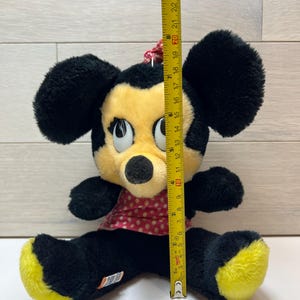 K&ouml;nnte beinhalten: Ein Vintage-Minnie-Mouse-Pl&uuml;schtier, ca. 23 cm gro&szlig;. Das Pl&uuml;schtier hat schwarzes Fell, ein gelbes Gesicht und gelbe F&uuml;&szlig;e sowie ein rotes Kleid mit Polka Dots. Das Spielzeug sitzt mit gespreizten Beinen.