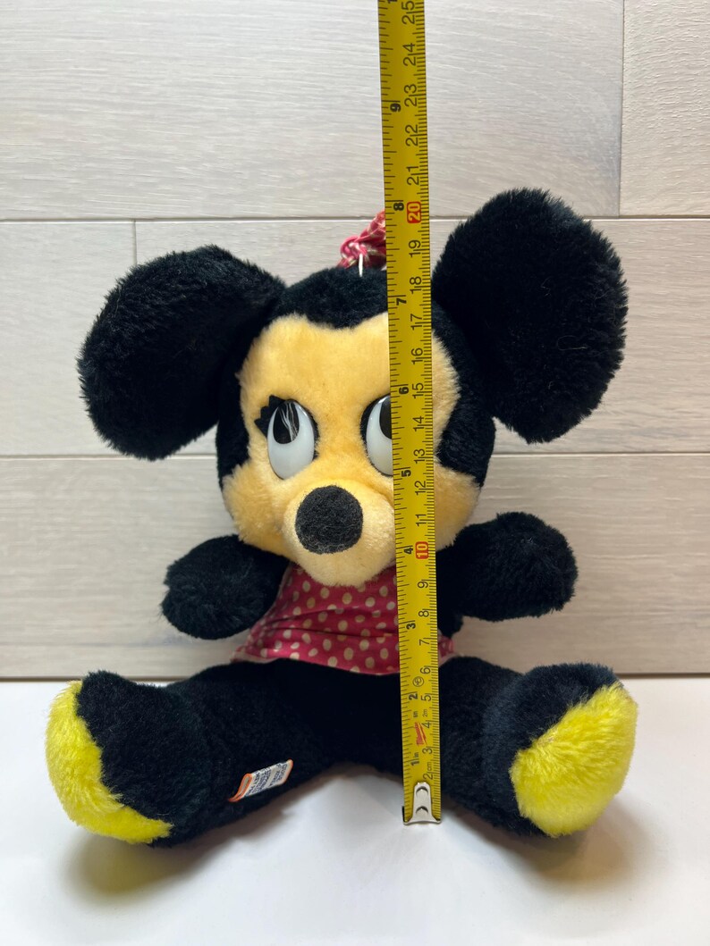 K&ouml;nnte beinhalten: Ein Vintage-Minnie-Mouse-Pl&uuml;schtier, ca. 23 cm gro&szlig;. Das Pl&uuml;schtier hat schwarzes Fell, ein gelbes Gesicht und gelbe F&uuml;&szlig;e sowie ein rotes Kleid mit Polka Dots. Das Spielzeug sitzt mit gespreizten Beinen.