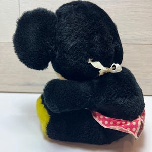K&ouml;nnte beinhalten: Ein Vintage-Pl&uuml;schtier in Schwarz mit gro&szlig;en Ohren und einem gelb-rosa Tupfenakzent. Das Spielzeug hat ein wei&szlig;es Band um den Hals gebunden. Das Pl&uuml;schtier sitzt auf einer wei&szlig;en Oberfl&auml;che.