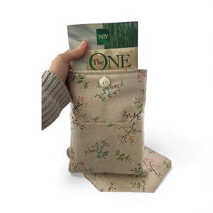 May include: A beige fabric book sleeve with a floral pattern, holding a book with the title "The ONE". The sleeve has a button closure and a front pocket. The floral design includes pink and green accents.
