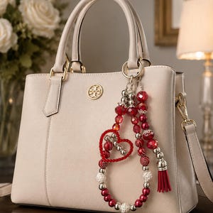 Puede incluir: Un bolso estructurado de color crema con dos asas superiores y un emblema con logotipo en tono dorado. Un dije decorativo cuelga del asa, con cuentas rojas y blancas, un marco rojo en forma de corazón y una pequeña borla roja en un anillo.