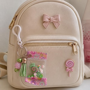 May include: A cream-colored mini backpack featuring a pink bow accent on the front pocket. The bag is decorated with a white plastic keychain holding green beads and a tassel, a clear pouch filled with miniature candy charms, and a pink lollipop pin.