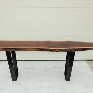 May include: A long, dark brown wooden table with a live edge top and black metal legs. The table is sitting on a gray concrete surface.