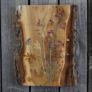 May include: A wooden slice with a natural bark edge, featuring pressed flowers and leaves in shades of pink, purple, and brown. The flowers are arranged in a vertical pattern, creating a rustic and elegant design.