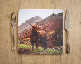 May include: A square placemat featuring a Highland cow standing in a mountainous landscape. The cow has long, shaggy brown fur and large horns. A fork and knife are placed on a wooden surface, framing the placemat.