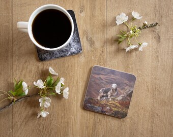 May include: A white ceramic mug filled with black coffee sits on a marble-patterned coaster. A second coaster features a sheep in a mountainous landscape. White flowers and branches are arranged on a wooden surface.
