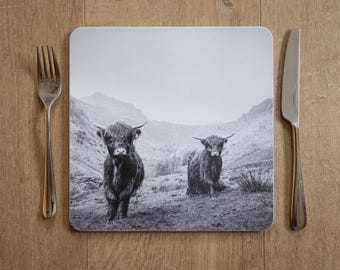 May include: A black and white placemat featuring two Highland cows in a mountainous landscape. A silver fork and knife are placed on either side of the placemat on a wooden surface. The placemat has rounded corners.