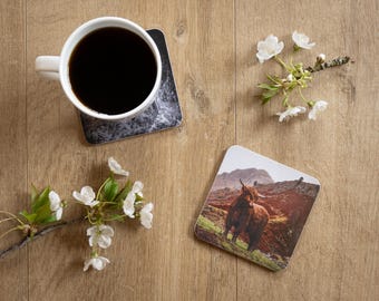 May include: A white coffee mug filled with black coffee sits on a marble-patterned coaster. A second coaster features a Highland cow in a landscape. White flowers and branches are arranged on a wooden surface.
