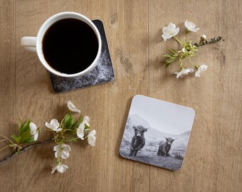 May include: A white coffee mug filled with black coffee sits on a square coaster with a marble design. A second coaster features a black and white image of two Highland cows. White flowers and branches are arranged on a wooden surface.