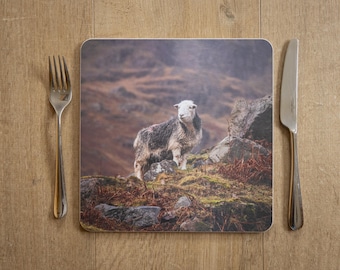 May include: A square placemat featuring a sheep standing on a rocky hillside. The sheep has a white face and gray and white wool. A fork and knife are on either side of the placemat, set on a wooden surface.