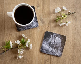 May include: A white coffee mug filled with dark liquid sits on a square coaster with a black and white marble design. A second coaster features a horse image. White flowers and a wooden surface complete the scene.