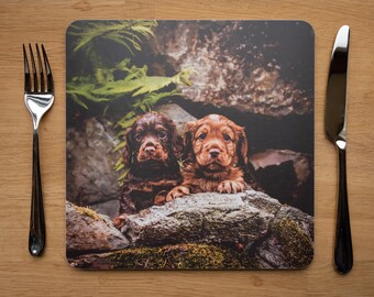 May include: Two brown and red puppies with floppy ears are peeking out from behind rocks. The puppies are looking directly at the camera.