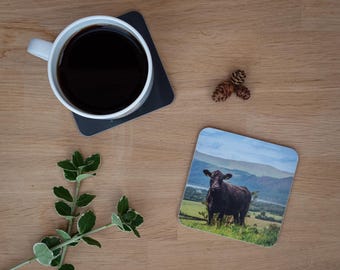 May include: A white mug filled with dark coffee sits on a dark gray square coaster. A second coaster features a black cow in a green field with mountains in the background. A sprig of green leaves and two pinecones are also on the wooden surface.