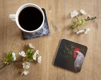 May include: A white coffee mug filled with dark liquid sits on a coaster with a marble design. A second coaster features a rooster image. White flowers and greenery are arranged on a wooden surface.